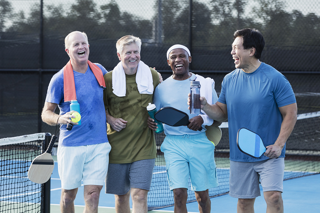 A multi-ethnic group of four mature and senior men walking off a pickleball court, laughing and conversing. They are taking a break after playing, towels around their shoulders, holding water bottles. The African-American man is the oldest, in his 70s. The others are in their 50s and 60s.